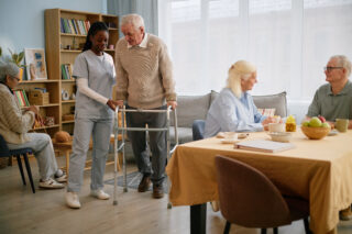 Elderly man using walker assisted by caregiver in care facility with other seniors in background. Room filled with books, chairs, and a table with breakfast items arranged